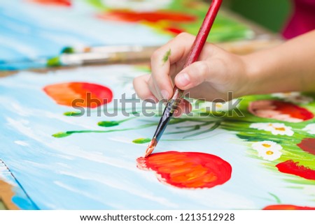 Child paints a picture by gouache. Kid drawing poppies and chamomiles. The hand and paint brush. Closeup, selective focus