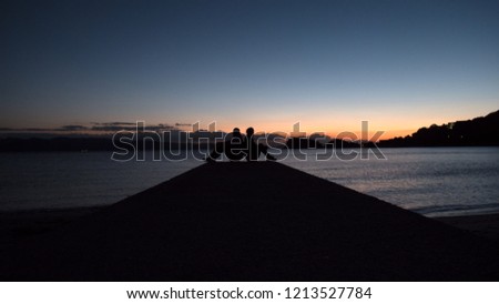 A couple sit back to back on a sea pier at sundown to create a beautiful romantic photo. The 2 people are silhouetted against a sunset sky and a calm ocean. 