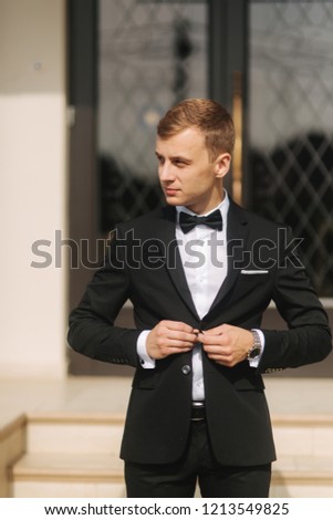 Fashion groom in trendy suit posing to photographer. Blond hair man with bow tie and other accsessories stand by the beautiful hotel