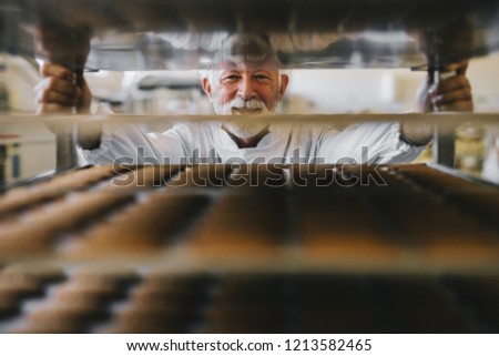 Picture of professional mature male baker man in white work uniform looking at the camera. Standing in front of the shelves full with fresh baked cookies.