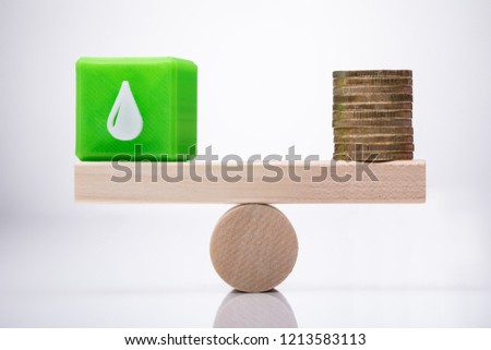 Green Cubic Block With Waterdrop Icon And Stacked Coins Balancing On Wooden Seesaw
