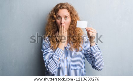 Young redhead woman over grey grunge wall holding blank visit card cover mouth with hand shocked with shame for mistake, expression of fear, scared in silence, secret concept