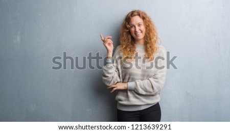 Young redhead woman over grey grunge wall with a big smile on face, pointing with hand and finger to the side looking at the camera.