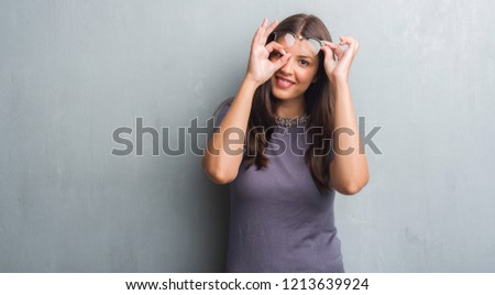Young brunette woman over grunge grey wall wearing glasses with happy face smiling doing ok sign with hand on eye looking through fingers