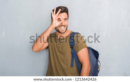 Handsome tourist young man over grey grunge wall wearing backpack doing ok gesture with hand smiling, eye looking through fingers with happy face.