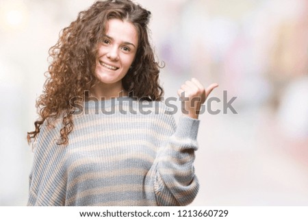 Beautiful brunette curly hair young girl wearing winter sweater over isolated background smiling with happy face looking and pointing to the side with thumb up.