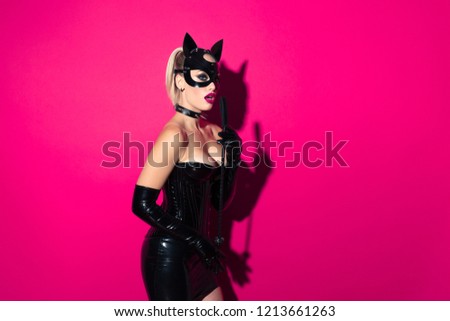 Beautiful dominant blonde vamp mistress bdsm girl with fashion makeup in glamour latex skirt, corset, collar and bdsm black leather fetish cat mask posing with whip on hot pink background