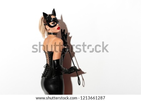 Beautiful dominant blonde vamp mistress bdsm girl with fashion makeup in glamour latex skirt, corset, collar and bdsm black leather fetish cat mask posing with whip on white background