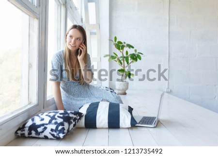 Charming young girl talking with smartphone looking out the window sitting in social networks using a laptop and wireless connection