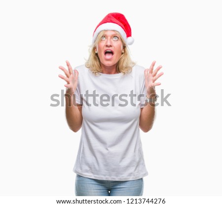 Middle age blonde woman wearing christmas hat over isolated background crazy and mad shouting and yelling with aggressive expression and arms raised. Frustration concept.