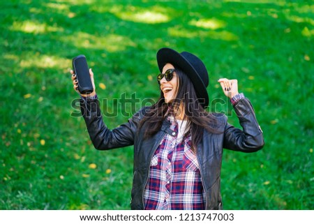 Happy mixed race young woman listening to music with wireless portable speaker. Hispanic hipster girl dancing to rhythm and singing along melody in the park