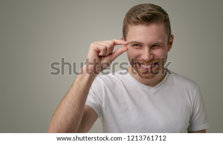 A handsome gay man has bristles, giggles at the camera and shows off something small, dressed casually, isolated on a white background.  A young man demonstrates a tiny thing in the room.