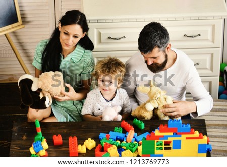 Parents and son with smiling faces make brick constructions. Mom, dad and boy play with toys on wooden background. Young family spends time in playroom. Love and family games concept