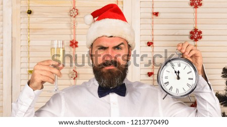 Santa Claus in red hat with angry face in festive room. Guy on wooden wall background with garlands. Celebration and New Year time concept. Man with beard holds glass of champagne and alarm clock