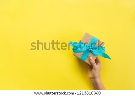 Woman hands give wrapped valentine or other holiday handmade present in paper with blue ribbon. Present box, decoration of gift on yellow table, top view with copy space.