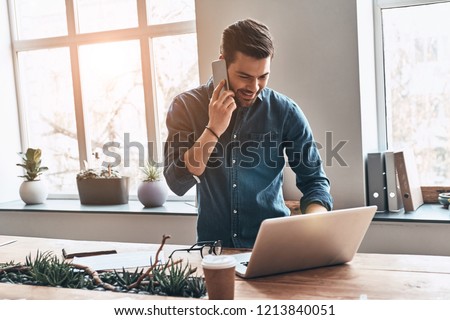 Good business talk. Handsome young man talking on smart phone and using computer while working in the office Royalty-Free Stock Photo #1213840051