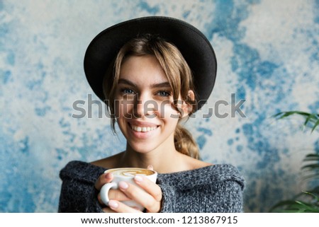 Close up portrait of a young woman dressed in sweater and hat over blue wall background, holding cup of hot coffee, looking at camera