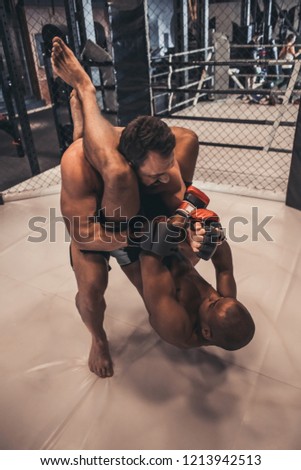 Two men in gloves and shorts are fighting in cage using grappling