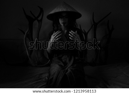 Young woman with scary make up, japanese (chinese) hat and deer horns sitting in dark room. Black and white. Halloween costume