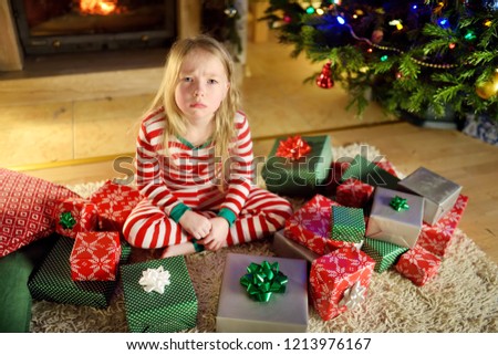 Cute little girl feeling unhappy with her Christmas gifts. Child sitting by a fireplace in a cozy dark living room on Xmas eve. Too many presents for Christmas.
