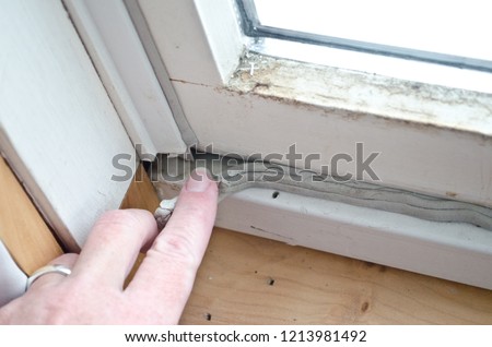 Caucasian woman's hand pressing in grey caulk in old window casement with bare wood windowsill for winterizing to keep out cold drafts and keep energy costs and heating bill down. Royalty-Free Stock Photo #1213981492