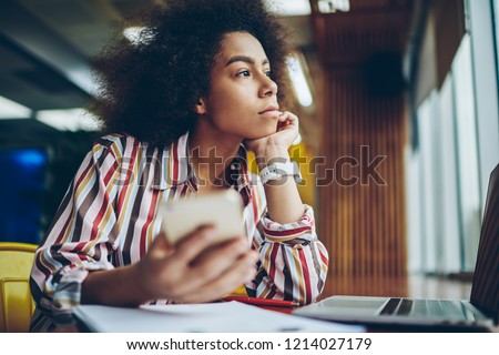 Pondering african american young woman thinking on creative ideas for publication in blog on smartphone device sitting in coffee shop.Thoughtful dark skinned hipster girl looking out of window