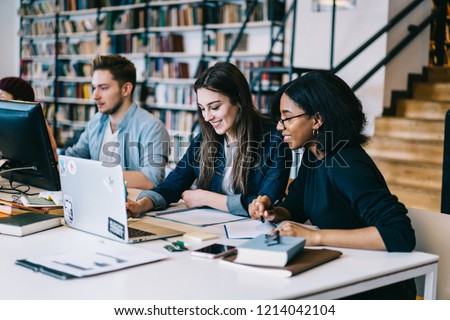 Cheerful diversity group of students sitting in university campus with books and technology learning for exams, multiracial hipster girls smiling and talking while making online research on netbook Royalty-Free Stock Photo #1214042104