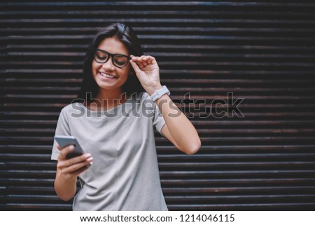Happy young hipster girl in eyewear holding modern smartphone and enjoying time near copy space area, cheerful female blogger with smile on face using cellular for chatting while browsing 4g internet