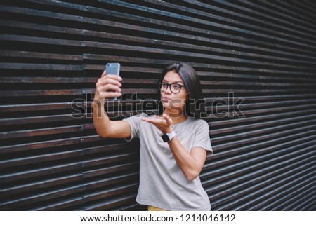 Attractive female blogger sending 'fly kiss' to followers while making new video blog using contemporary smartphone and standing near copy space area for advertising text and messages outdoors