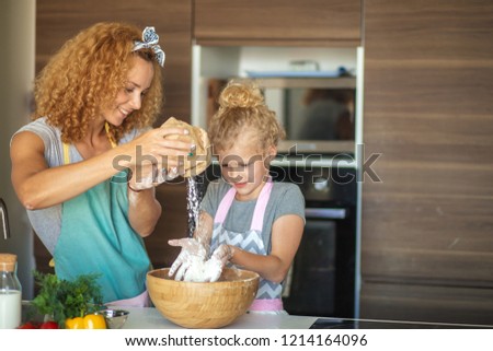 Pretty female housewife cooker wears apron and headband on head, has glad cheerful expression while demonstrating little blond girl how to make cake, Home Cooking, Homemade Food