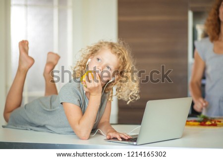 Preschool girl with blonde curly hair eating paprica and watching cartoons using laptop and earphones in the room