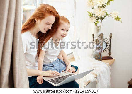 Red haired attractive babysitter entertaining her kid under supervision, sitting on window sill and watching cartoons on laptop.
