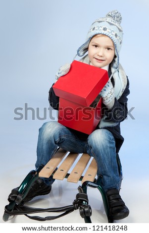 Happy Smiling Boy With A Big Red Gift Sitting On A Sled Over Blue Background