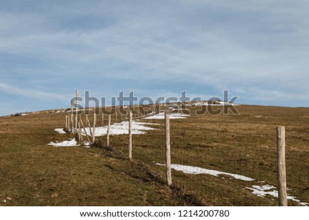 Wooden fence posts and barbed wire marking the property line of a prairie with dried grass and patches of snow under blue sky with white clouds