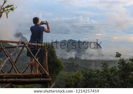 Female tourist standing on the wooden platform and using mobile phone taking picture of mist over the mountain at early morning light in Krabi Province, Southern of Thailand
