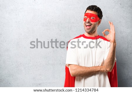 Superhero man with mask and red cape showing an ok sign with fingers while winking an eye on textured grey background