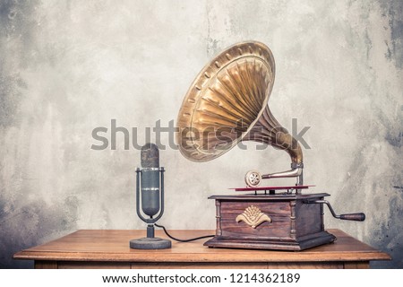 Vintage antique gramophone phonograph turntable with brass horn and big aged studio microphone on wooden table front concrete wall background. Retro old style filtered photo Royalty-Free Stock Photo #1214362189
