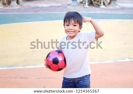 Sport and kid concept, At the basketball court, a funny child boy holding a football. He wear white shirt. He showing hand biceps muscles strength.