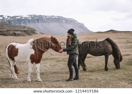 Woman in black hat, jeans and green jacket feeds red horse with white spots in the icelandic field with mountains on the background