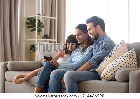 Married diverse couple spend free time on weekend with small daughter sitting on sofa in living room at home. Positive mom daddy little kid use smartphone watching cartoons videos online having fun