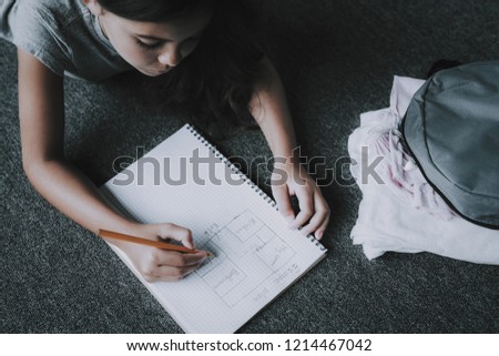 Top View of Girl Draws in Notepad Lying on Floor. Closeup Portrait of Black-Haired Child Lying on Gray Carpet and Writes with Pencil in White Notebook. Leisure Time Concept