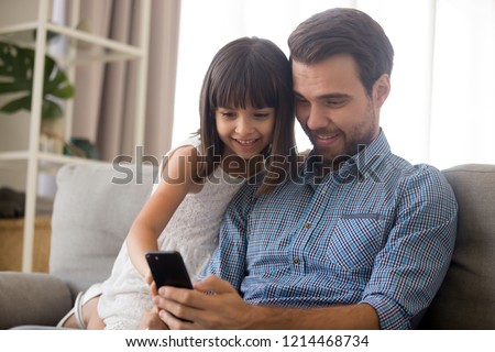 Cheerful positive multi-ethnic family sitting on couch in living room at home Caucasian father daughter smiling looking on smartphone screen have fun watching cartoons chatting playing games