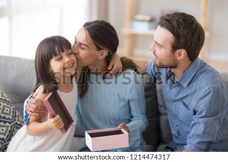 Diverse young family sitting on couch in living room at home grateful mom kissing on cheek little preschool daughter. Loving husband, kid prepare for mum wife a gift at birthday or mother day holiday