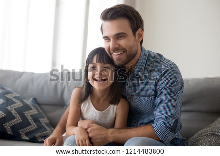 Laughing diverse family sitting on couch in living room at home. Young father little daughter having video call using laptop or watching movie comedy or cartoon. Weekend activities free time concept