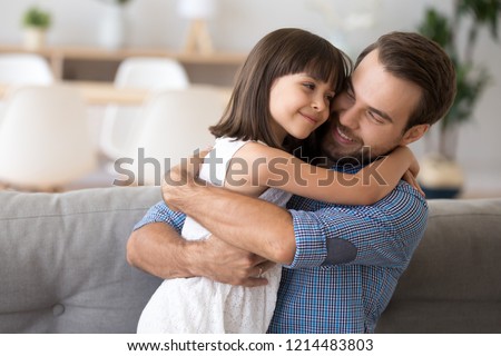 Adorable preschool daughter wearing white dress hugging loving father looking at him with love and tenderness. Multi-ethnic diverse friendly family on couch at modern home spending free time together Royalty-Free Stock Photo #1214483803