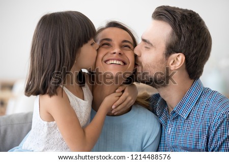 Positive smiling multi-ethnic people sitting together on couch at home. Little preschool daughter hugging kissing mother. Loving husband kiss on cheek wife. Happy friendly multiracial family concept