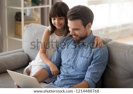 Positive multi-ethnic family father and little daughter spend free time on weekend sitting on couch at home watching funny videos cartoons browsing internet or have video call using wireless computer