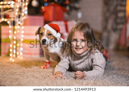 Merry Christmas. Little girl child at home at the fireplace with a dog Jack Russell Terrier and a New Year tree with gifts and luminous garlands celebrates Christmas