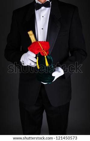 Gentleman wearing a tuxedo holding a champagne bottle wrapped up in a festive holiday gift bag. Man is unrecognizable on a light to dark gray background.