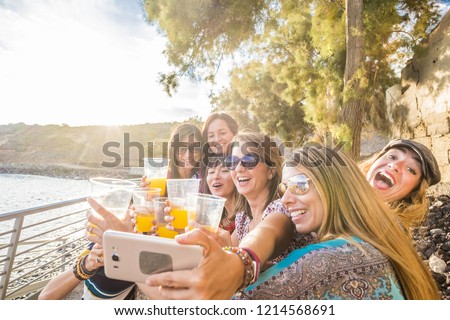 Group of women people crazy taking a selfie picture all together with a smartphone while celebrate with orange healthy juice freuit outdoor near the beach in summer time. fun and happiness for friends